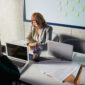 Managing director sitting in front of whiteboard with green marker and looking at another person at her desk